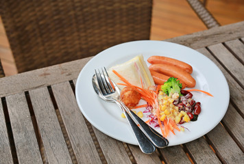 Breakfast with sandwich, sausage and salad in a white dish on wooden table.