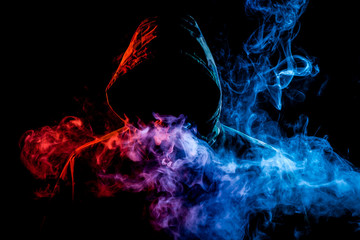 A young man in a black hood smokes a vape and exhales a colored blue, pink, yellow smoke on a black...