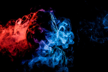 A young man in a black hood smokes a vape and exhales a colored blue, and red smoke on a black isolated background