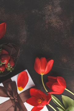 Holiday dark background. White gift box, red tulips and chocolate-strawberry dessert. Mother's Day / St. Valentine's Day / birthday concept.