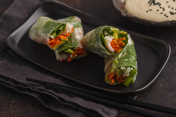 Fresh handmade vegan asian spring rolls with rice noodles, avocado, carrots and tahini dressing on black dish, dark background.