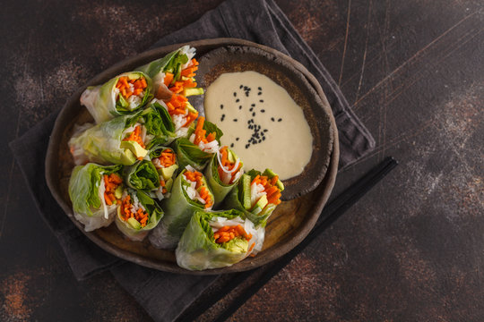Fresh handmade vegan asian spring rolls with rice noodles, avocado, carrots and tahini dressing on black dish, dark background. Top view, copy space.