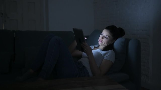 lateral pan shot of young beautiful happy and relaxed latin woman lying on home couch late night using digital device laptop tablet pad watching internet movie 