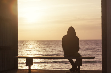 Silhouette of a man in a hooded jacket sitting on a bench and watching the sunset. Loneliness, sadness, alone with nature