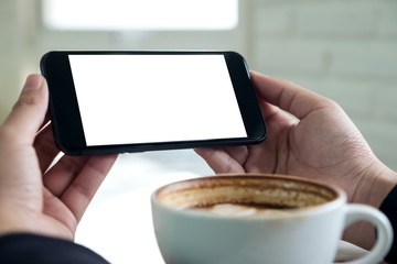 Mockup image of hands holding black mobile phone with blank white screen for watching and playing games with a cup of coffee in cafe