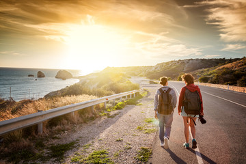 Two young female travel girlfriends walking along a highway, against a background of sunset and sea...