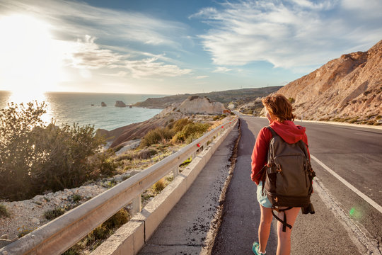 A young woman traveler walks along a highway, against the background of a sunset and sea coast. Travel and freedom, adventures and directions for travel
