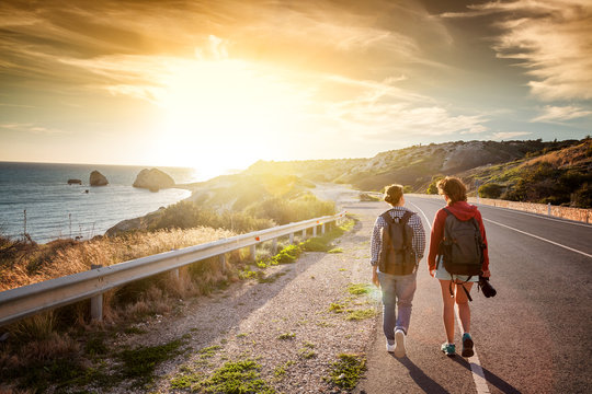Two young female travel girlfriends walking along a highway, against a background of sunset and sea coast. Travel and freedom, adventures and directions for travel