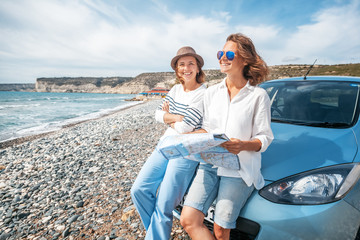 Two young beautiful female friends travel together by car, look at the road map against the sea,...
