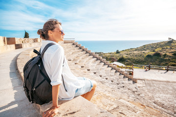 A young beautiful woman traveler sits on the steps of an amphitheater in an architectural antique museum overlooking the sea. A trip to antiquity, Cyprus, Park Kourion