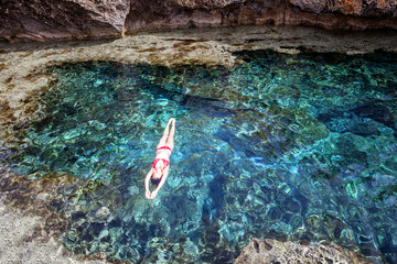 Fototapeta na wymiar Attractive young woman in red bikini swims meditating in a bay with turquoise water surrounded by caves, shot from above