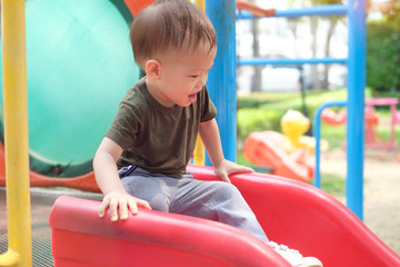 Fototapeta na wymiar Cute smiling little Asian 18 months / 1 year old toddler baby boy child playing on a slide in playground on summer sunny day, Kid Sliding Down Slide, Free play Healthy Child Development concept
