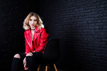 Studio portrait of blonde girl in red leather jacket posed on chair against brick wall.
