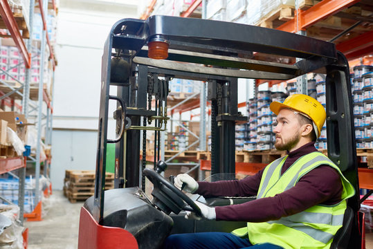Side view portrait of young warehouse worker sitting inside forklift moving goods from tall storage shelves, copy space