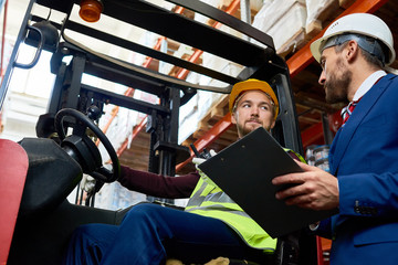 Low angle portrait of mature businessman talking to warehouse worker sitting in forklift car, copy space © Seventyfour