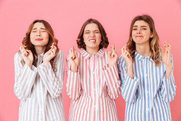 Portrait of three joyful young girls 20s wearing colorful striped pajamas with closed eyes willing good luck keeping fingers crossed during sleepover, isolated over pink background