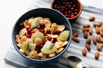 Delicious crispy cornflakes with kiwi pieces and cranberries in bowl, almonds on dish cloth, closeup, healthy breakfast