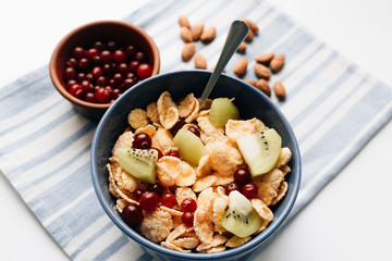 Delicious crispy cornflakes with kiwi pieces and cranberries in bowl, almonds on dish cloth, closeup, healthy breakfast