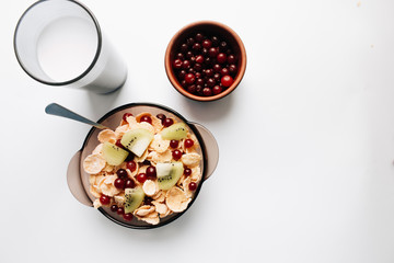Delicious crispy cornflakes with kiwi pieces and cranberries in bowl, glass of milk on white background, closeup, healthy breakfast