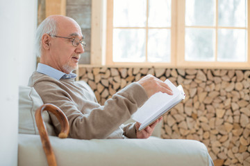 Interesting book. Busy charming senior man sitting on couch while holding book and reading