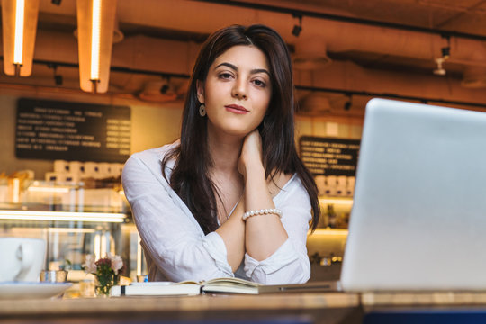 Portrait of young businesswoman, student, dressed in white blouse, sitting at table in cafe in front of computer.