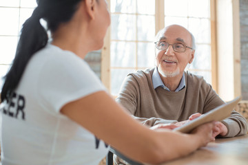 Twisted plot. Cheerful merry senior man laughing while listening to volunteer and sitting on blurred background
