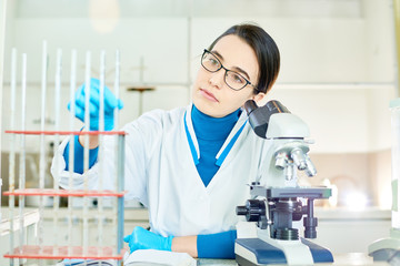 Portrait of pretty young medical technician wearing white coat and rubber gloves sitting at laboratory bench and taking graduated pipette from holder in order to study its content with microscope.