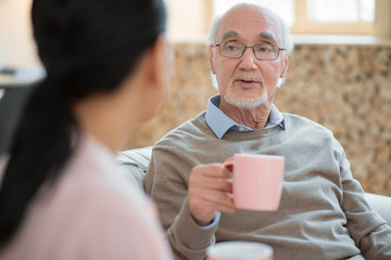 Cosy atmosphere. Attractive smart senior man chatting with caregiver while drinking tea and holding mug