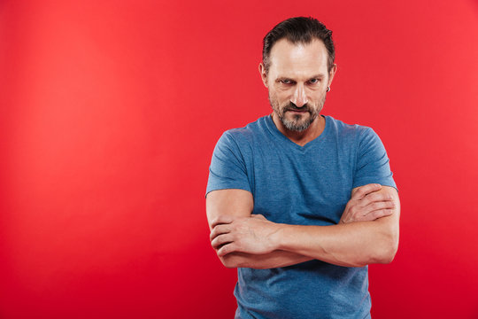 Photo of aggressive man wearing casual t-shirt standing with hands crossed and looking on camera with angry gaze, isolated over red background