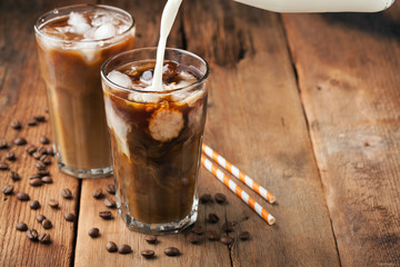 Ice coffee in a tall glass with cream poured over and coffee beans on a old rustic wooden table. Cold summer drink on a dark wooden background with copy space