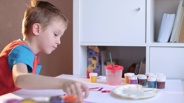 The boy draws with pencils and paints on a white sheet. near a lot of colored jars with paint. Gouache, watercolor theme.