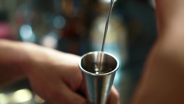 Young bartender pours tequila into glass on counter in trendy bar. Man fills glass with alcoholic liquid from bottle and gives it to client indoors. He holds metal object in arm and pours drink with