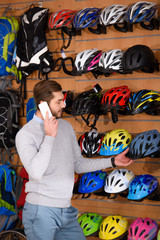 Fototapeta na wymiar smiling young man talking by smartphone and choosing bicycle helmets in bike shop