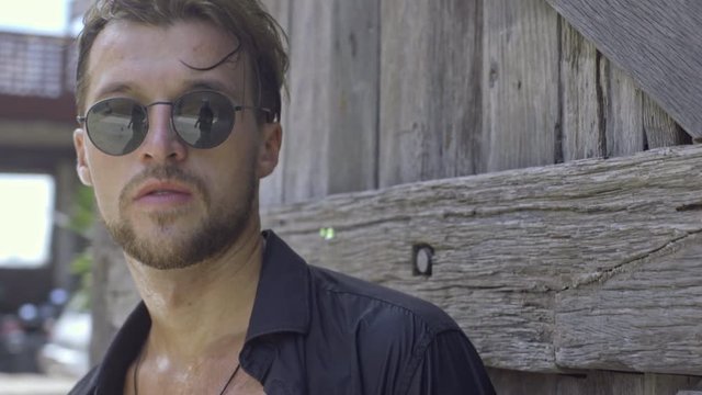 Tilt down shot of handsome young man in sunglasses and unbuttoned shirt standing with longboard near wooden wall at hot summer day