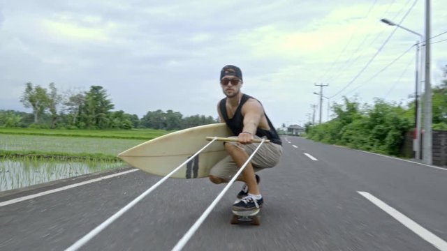 Young skateboarder in sunglasses holding surfboard and skitching after scooter along tropical road in Bali