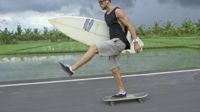 Young man in sunglasses riding skateboard along tropical road, holding surfboard and listening to music with headphones