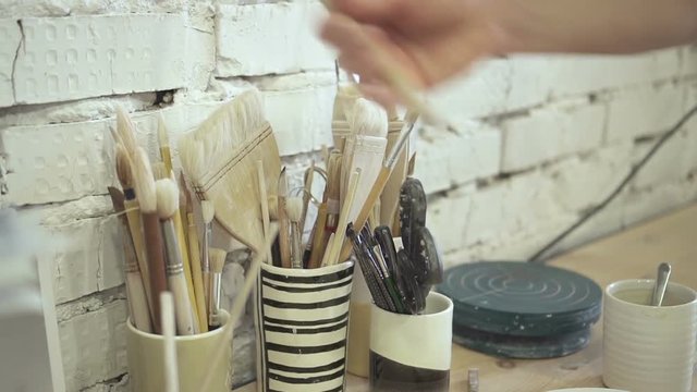 Male artist is taking brush for painting in pottery workshop. Young professional takes out tassel from jar on table in vintage studio indoors. Master chooses then takes fluffy brush for drawing. In