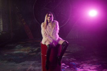 Young attractive woman with stylish clothes. Beautiful girl in fluffy pink fur coat sits on barrel. Neon light