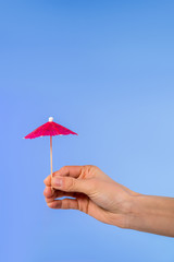 cropped view of female hand with cocktail umbrella, isolated on blue