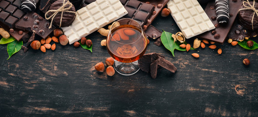 A glass of cognac, brandy, whiskey and a set of chocolate with cookies and sweets. On a black wooden background. Copy space for text. - obrazy, fototapety, plakaty