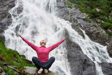 Caucasian female tourist sits with her arms wide open and looks up with delight against background of waterfall Balea in Romania. Rocky waterfall in the mountains.