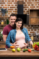 Fototapeta premium happy young pregnant couple hugging and smiling at camera while cooking together