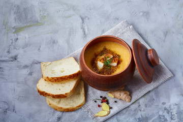 Clay pot of pumpkin soup on napkin over white textured background, close-up, selective focus, top view.