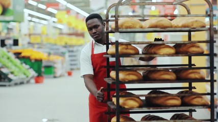 Close up handsome young african american man seller puts the bread on the counter in supermarket eating job occupation retail work baguette basket bake baker slow motion