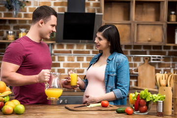 happy young pregnant couple holding glass and jug of fresh orange juice and smiling each other at kitchen