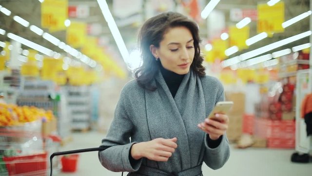 Attractive young woman walk checking to do list on phone in supermarket smiling shopping internet buying technology hypermarket shop mall smartphone shopper grocery market, girl food customer