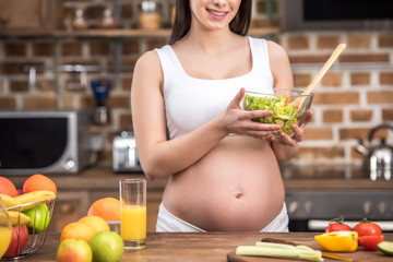 cropped shot of smiling young pregnant woman holding glass bowl with healthy vegetable salad in kitchen