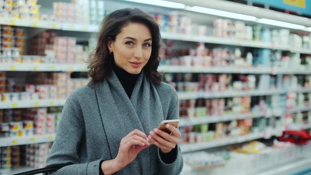 Beautiful young woman use phone in supermarket smiling look at camera shopping internet buying technology hypermarket shop mall smartphone shopper grocery market girl food customer slow motion