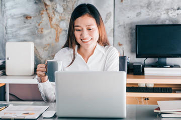 Asian businesswoman take a coffee break after working at laptop computer on desk with smiling...