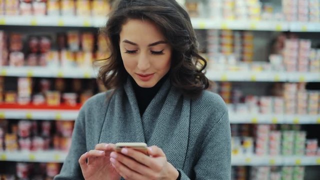 Happy young woman use phone in supermarket smiling look at camera shopping internet buying technology hypermarket shop mall smartphone shopper grocery market girl food customer slow motion